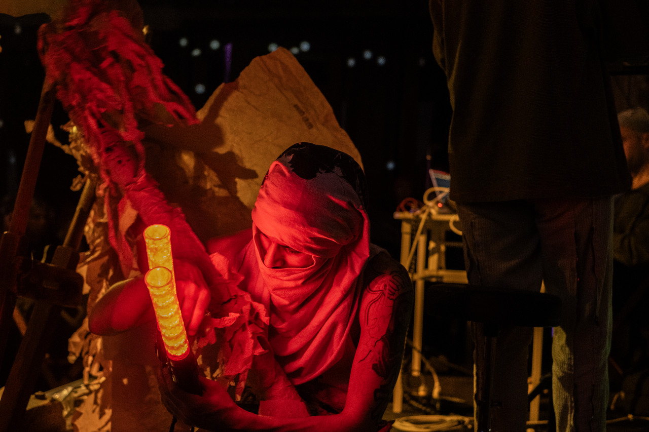Close-up color image of a performer holding red lights with a shirt wrapped around their head. Behind them, in the left side of the frame, a handmade paper costume is out of focus. On the right side of the frame there are two performers, one of them is standing with their back to the camera, and the other is sitting down at a table.
