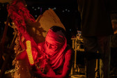 Close-up color image of a performer holding red lights with a shirt wrapped around their head. Behind them, in the left side of the frame, a handmade paper costume is out of focus. On the right side of the frame there are two performers, one of them is standing with their back to the camera, and the other is sitting down at a table.