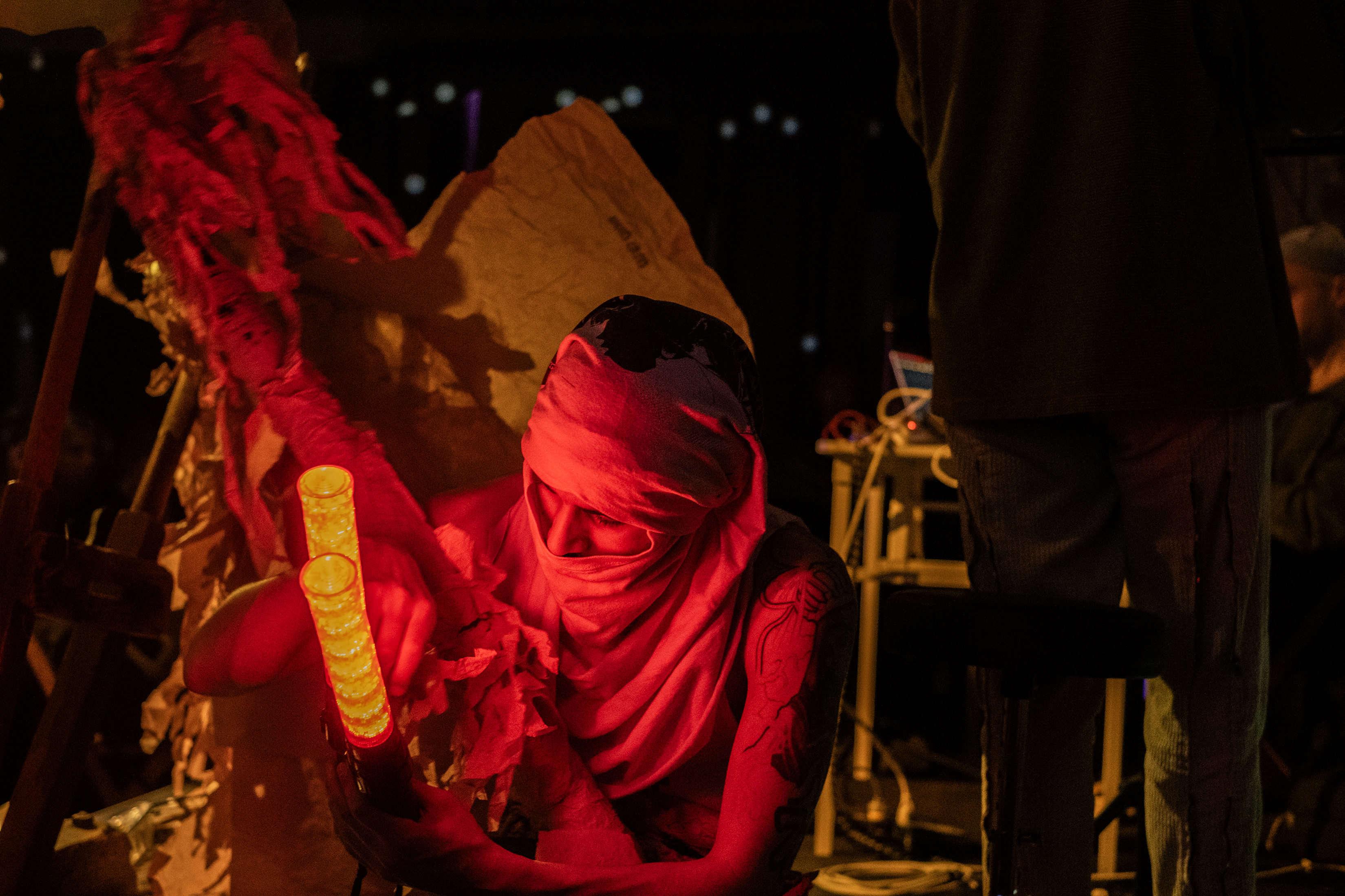 Close-up color image of a performer holding red lights with a shirt wrapped around their head. Behind them, in the left side of the frame, a handmade paper costume is out of focus. On the right side of the frame there are two performers, one of them is standing with their back to the camera, and the other is sitting down at a table.