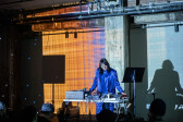 Color image of a performer on stage, standing in front of various types of electronic audio equipment. They are wearing a monochromatic purple outfit and glasses. The wall behind them is decorated with blue, orange, and purple projections. The left side of the room is lit with small blue dots reflecting from a disco ball. A few audience members are visible in the bottom edge of the image.