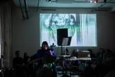 Color image of a performer on stage playing the cello. They are facing sideways to the audience, wearing sunglasses and a purple jacket. To their side there is a collection of audio equipment, as well as a music stand and a speaker. The audience is sitting down facing the stage. The room is dark, except for a projected image of a disco ball on the back wall.