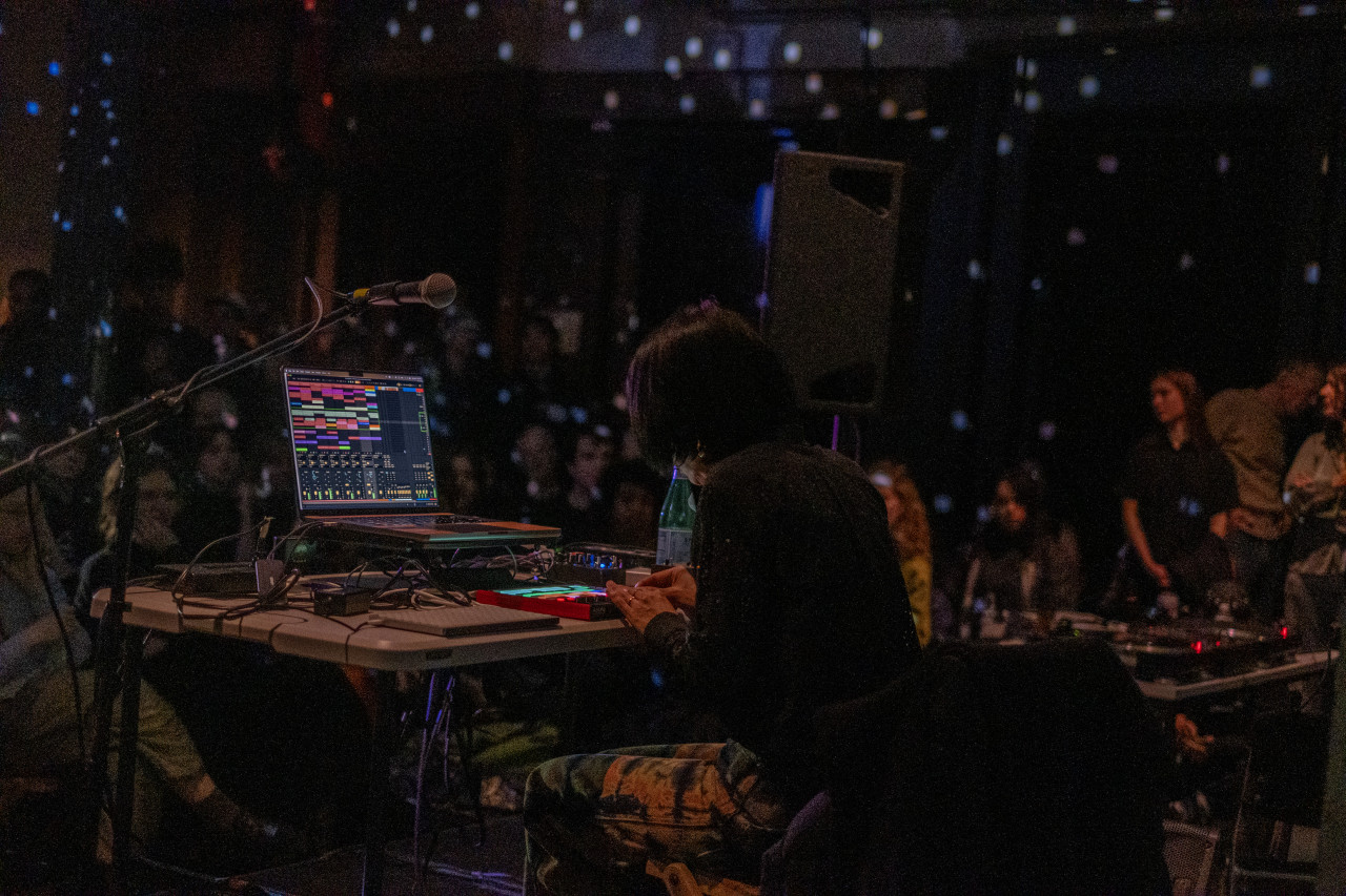Color image of a performer sitting on stage in front of a table with a laptop and audio equipment. They are leaning forward, using both hands on the machine. The laptop on the table shows live audio tracks, with bars of various colors. The room is dark and the audience is out of focus in the background. White dots of light fill the room.