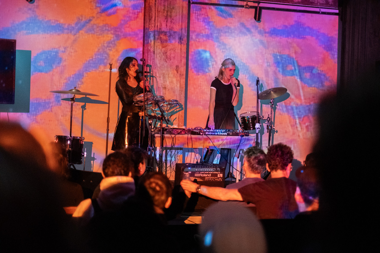 Color image of two performers on standing on stage. They are both looking to the side with microphones close to their mouths. They are surrounded by drums, cymbals, and a audio board on a table. The wall behind the performers is decorated with an abstract orange, blue, and pink projection, which also covers the performers. Multiple rows of seated audience members are visible in the foreground.