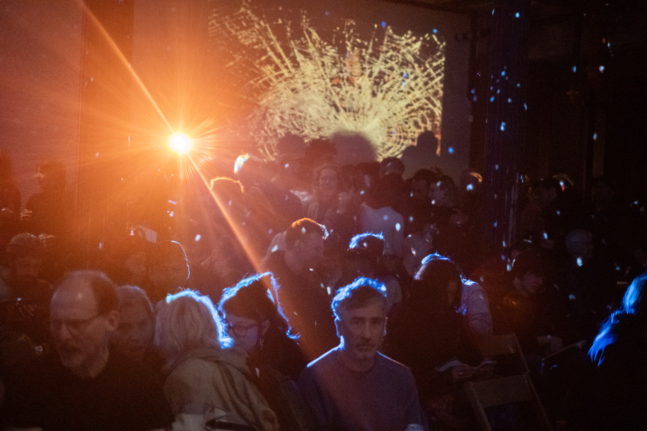 Color image of an audience in a room. Some of them are seated, others in the background are standing up. A bright yellow light is pointing into the camera, creating a dramatic light flare. The wall in the background features an abstract image projection, resembling a piece of shattered glass. The atmosphere in the room is hazy, and white dots of light from a disco ball fill the room.