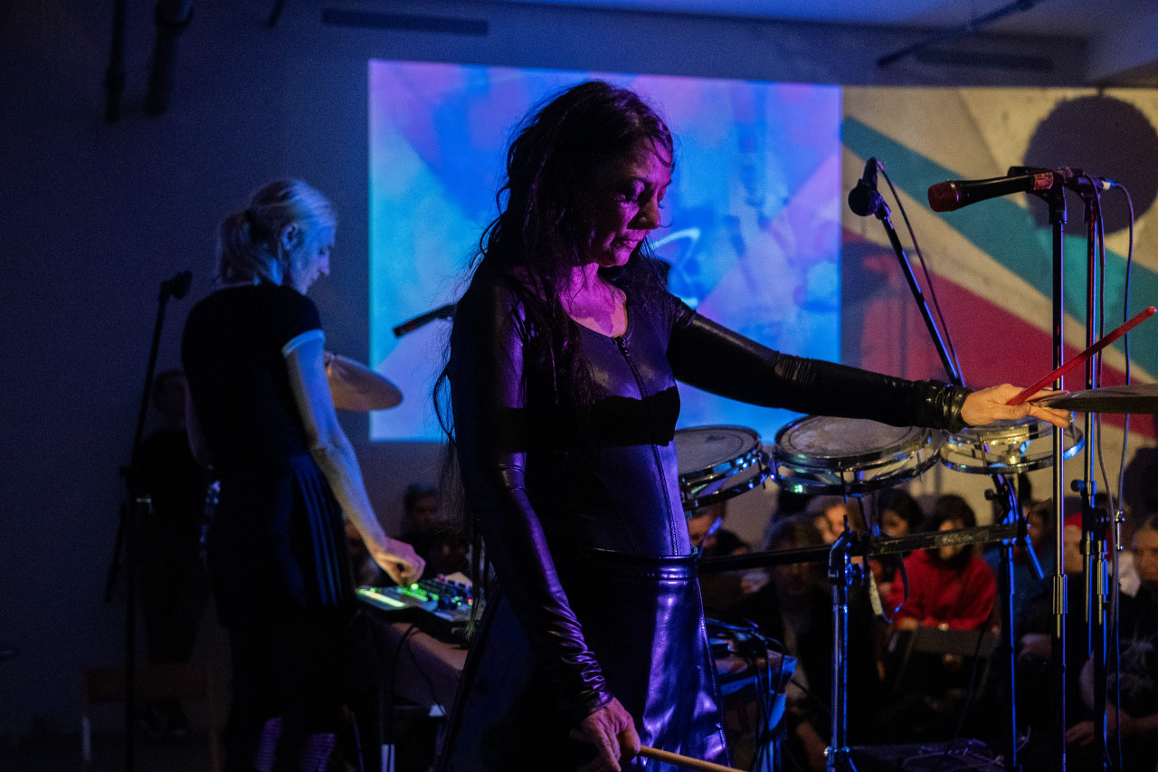 Color image of two performers on standing on stage. The performer in the foreground is lit with pink light. They are holding their arm out touching a cymbal with a red drum stock in hand. They are looking down with their eyes closed. The performer in the background is out of focus, they are bathed in blue light. Drums surround the stage. A seated audience is out of focus in the background. The back wall features two projections, one is an abstract arrangement of shades of blue. The other has sharp geometric shapes in red, green, and yellow.