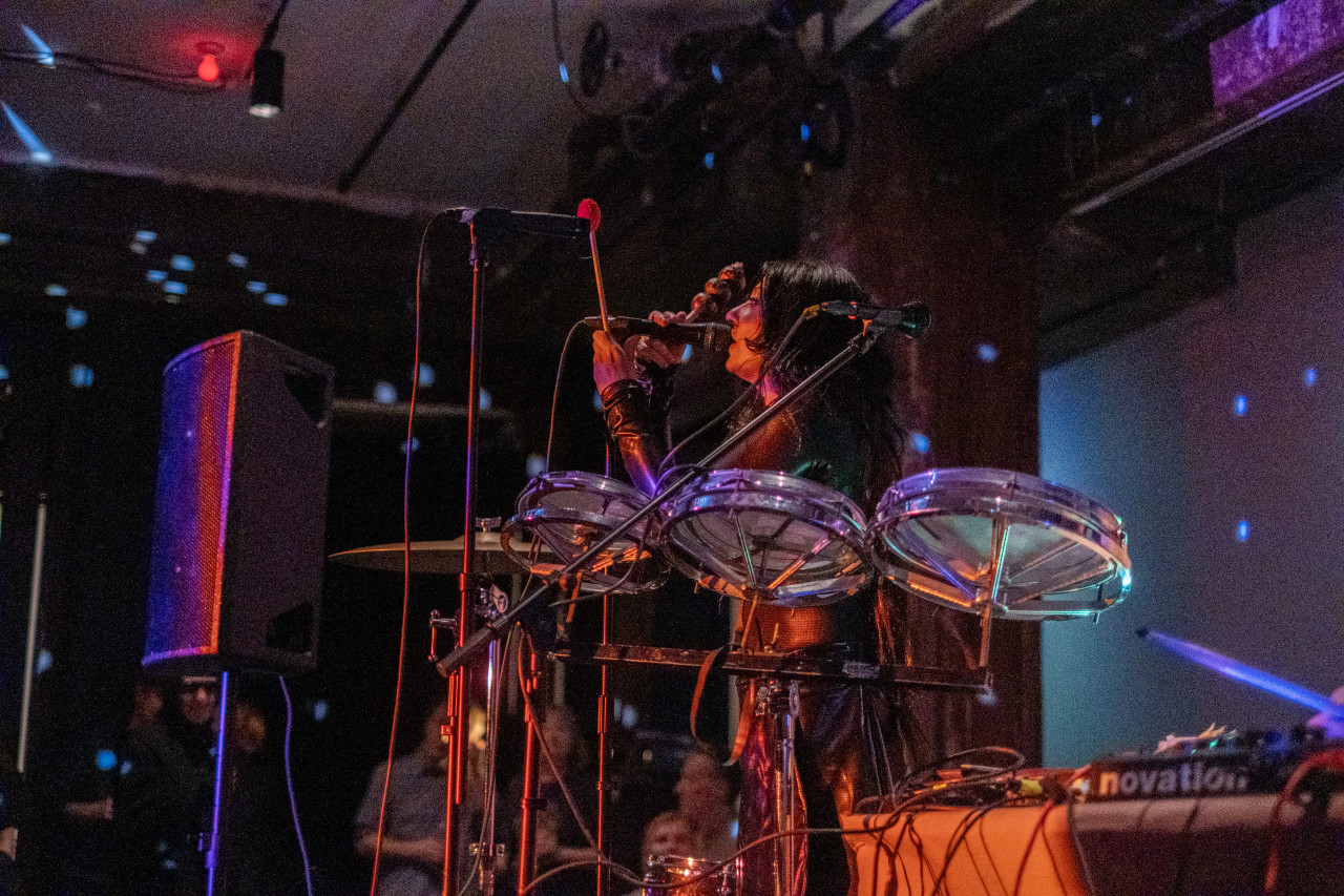 Color image of a performer on stage holding a microphone close to their mouth. They are holding a drum stick in one hand, standing next to a cymbal and a drum set on a stand. They are bathed in red and pink light, and the background is lit with blue, red, and purple light. Small white dots of light from a disco ball fill the room.