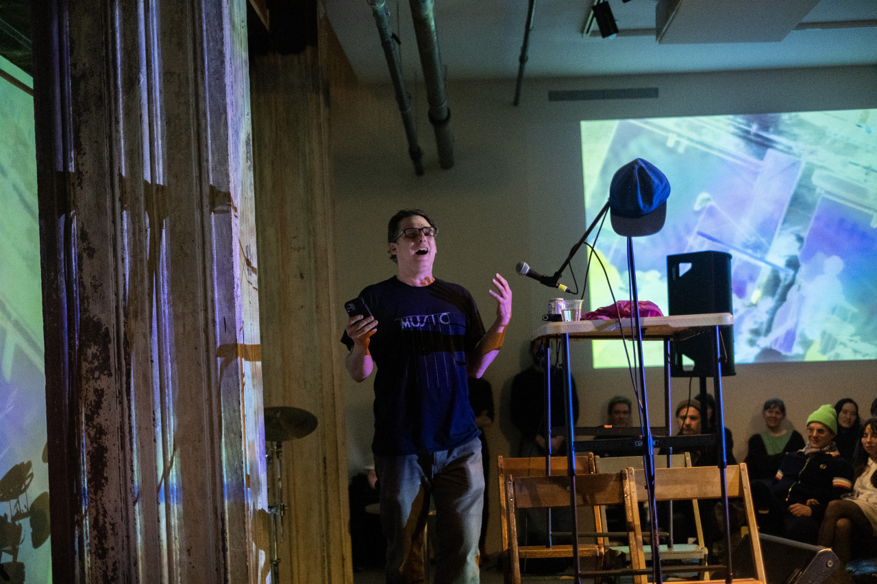 Color image of a performer on stage, standing sideways, with an audience facing them. The performer has their hands up and their mouth open in an exasperated manner. They are holding a phone in one hand. They are standing near a microphone on a stand, with a hat balanced on it. The audience is seated. The back wall features a distorted projection of a live feed of the performance.