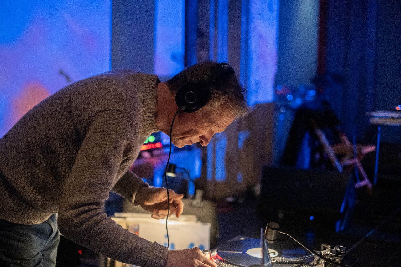 Color image of a performer leaning over a record player with headphones on. One of their hands is partly out of frame while touching the machine. The background is out of focus, and bathed in bright blue light. A small portable light is placed on top of the record player, illuminating both it and the performer.