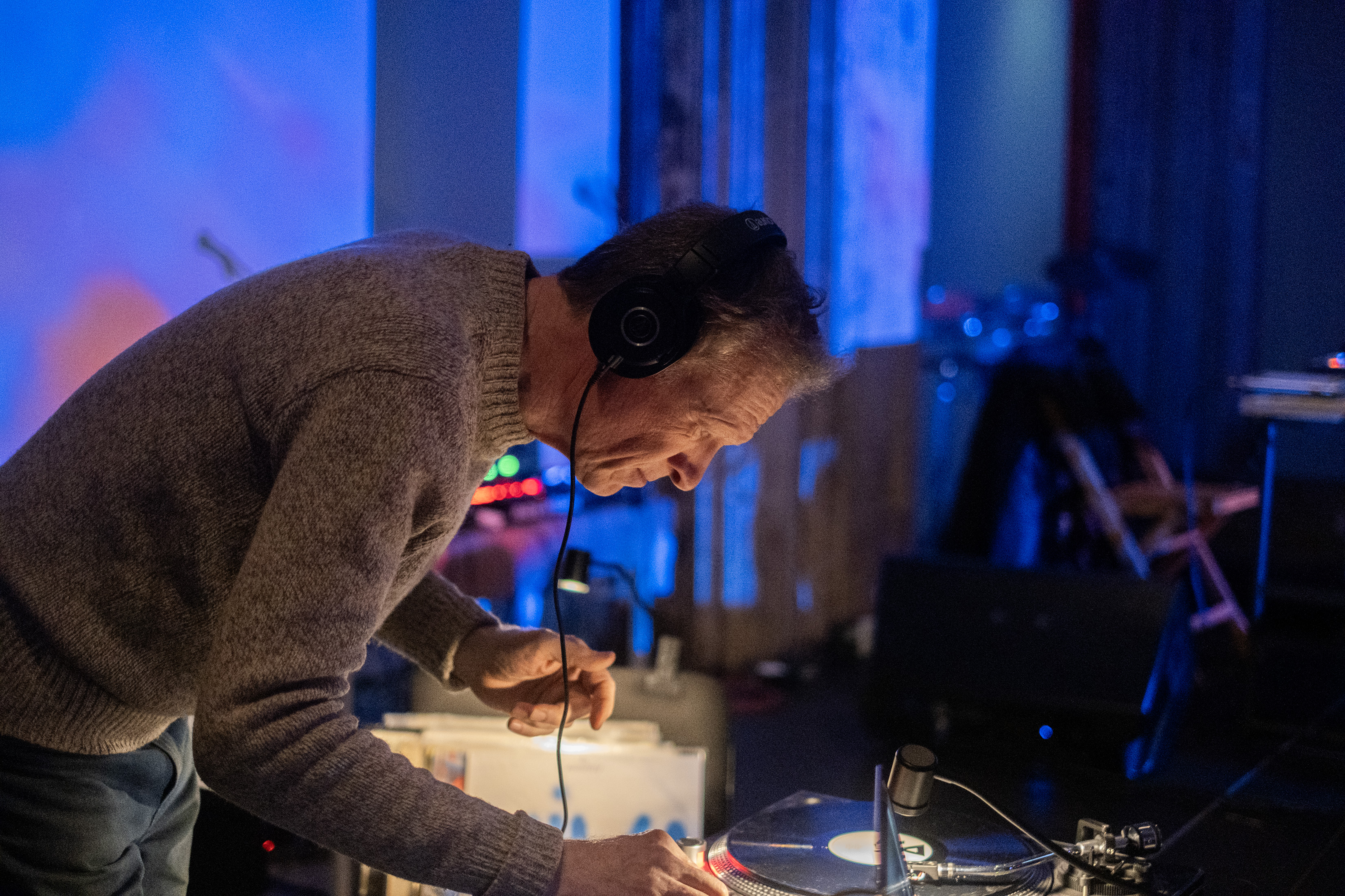 Color image of a performer leaning over a record player with headphones on. One of their hands is partly out of frame while touching the machine. The background is out of focus, and bathed in bright blue light. A small portable light is placed on top of the record player, illuminating both it and the performer. 