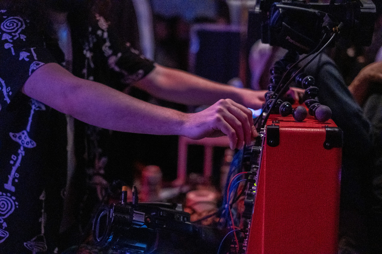 Close-up color image of a persons hands on a red audio mixing device. Various blue and red cords stick out of the machine. A camera on a tripod sits on top of the machine. The background is blurred, with a few audience members visible. The scene is lit with soft pink light.