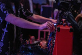 Close-up color image of a persons hands on a red audio mixing device. Various blue and red cords stick out of the machine. A camera on a tripod sits on top of the machine. The background is blurred, with a few audience members visible. The scene is lit with soft pink light.