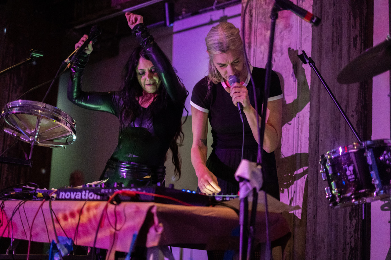 Color image of two performers on stage standing next to each other. The performer on the right is holding a microphone close to their mouth as they touch an audio board. The performer on the left is holding a microphone with both arms in the air, looking down at the board. There are drums to the sides of both performers. The scene is lit with soft pink and green light.