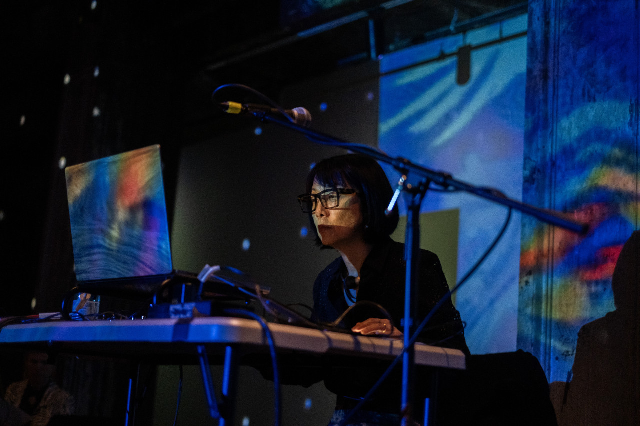 Close-up color image of a performer sitting on stage facing a laptop. The room is dark, lit with blue light and patches of rainbow abstract projections. There are white dots on the back walls reflecting from a disco ball. The performers face is lit up, they are wearing glasses and their hands are on the table.