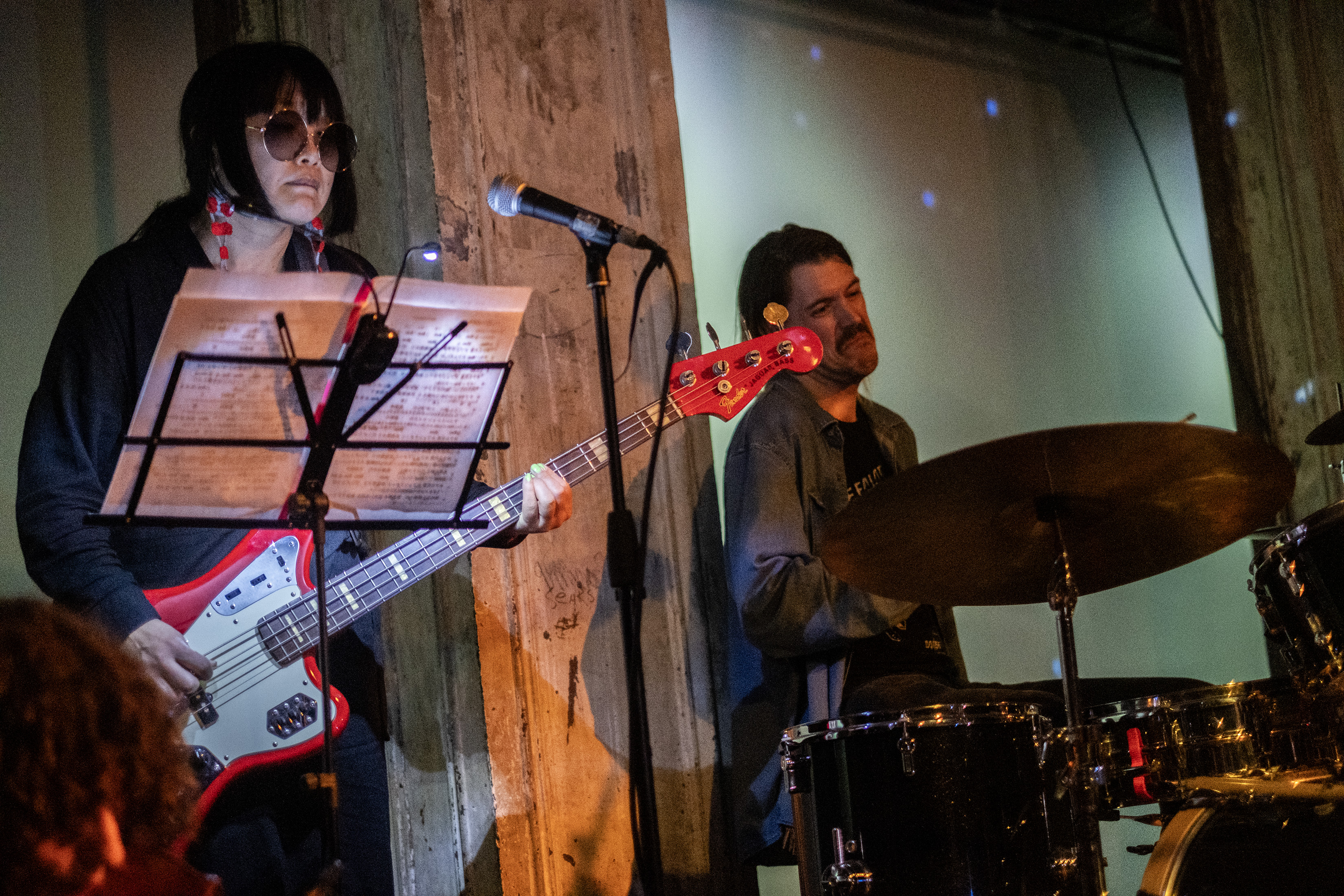 Color image of two performers on stage playing the guitar and drums. The performer on the left is wearing round sunglasses with a music stand in front of them. The back wall is lit with small white dots from a disco ball.