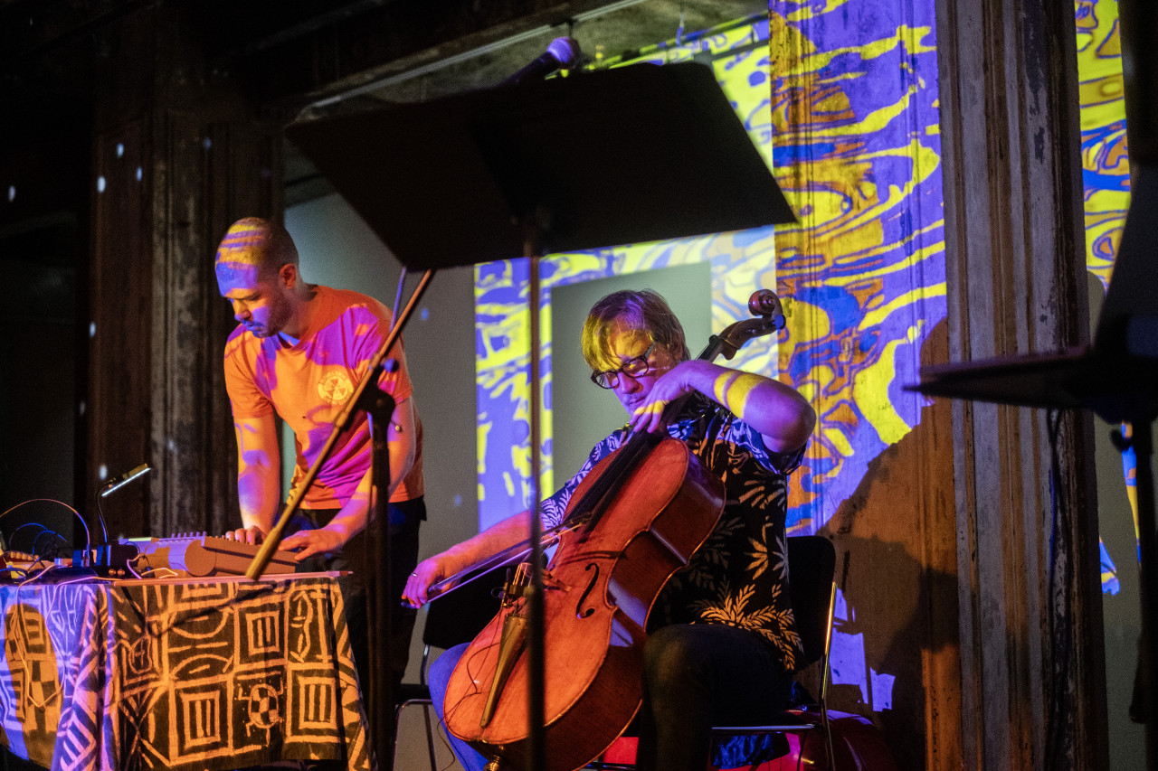 Color image of two performers, the performer on the left is standing over a 4-track tape teck on a table, the performer on the right is sitting playing the cello. A mic stand in the foreground splits the frame. A blue and yellow water-like image is projected on the wall.