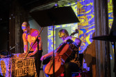 Color image of two performers, the performer on the left is standing over a 4-track tape teck on a table, the performer on the right is sitting playing the cello. A mic stand in the foreground splits the frame. A blue and yellow water-like image is projected on the wall.