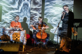 Color image of three performers on stage. The performer on the left is sitting at a table with a tape deck, the performer in the middle is playing cello with a bow, and the rightmost performer is standing with a microphone. One audience member is visible in the foreground. The musicians, as well as the wall behind them, are bathed in an abstract light projection.