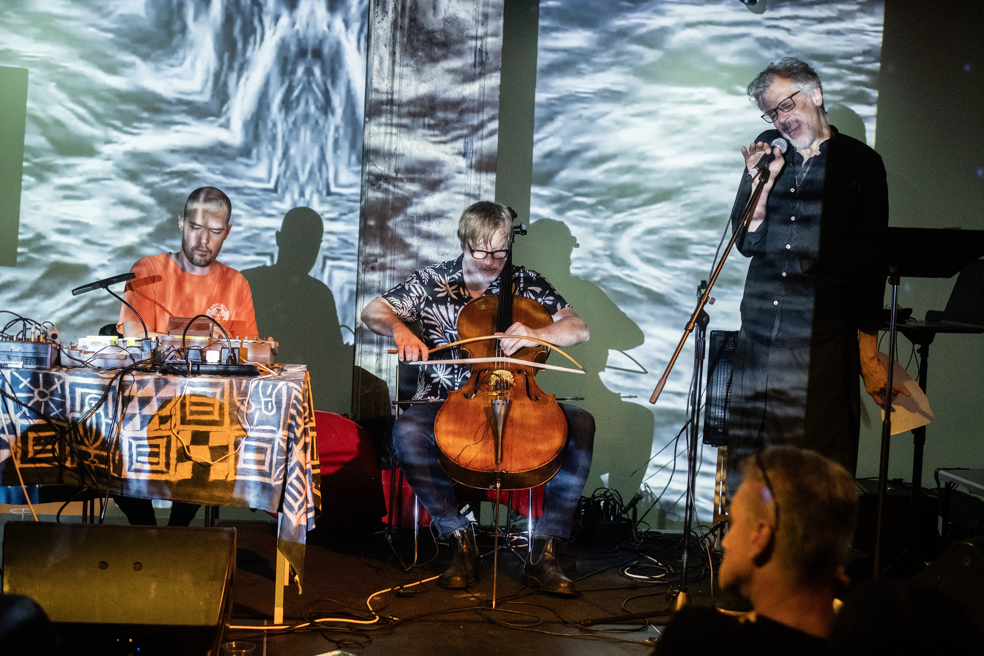 Color image of three performers on stage. The performer on the left is sitting at a table with a tape deck, the performer in the middle is playing cello with a bow, and the rightmost performer is standing with a microphone. One audience member is visible in the foreground. The musicians, as well as the wall behind them, are bathed in an abstract light projection.