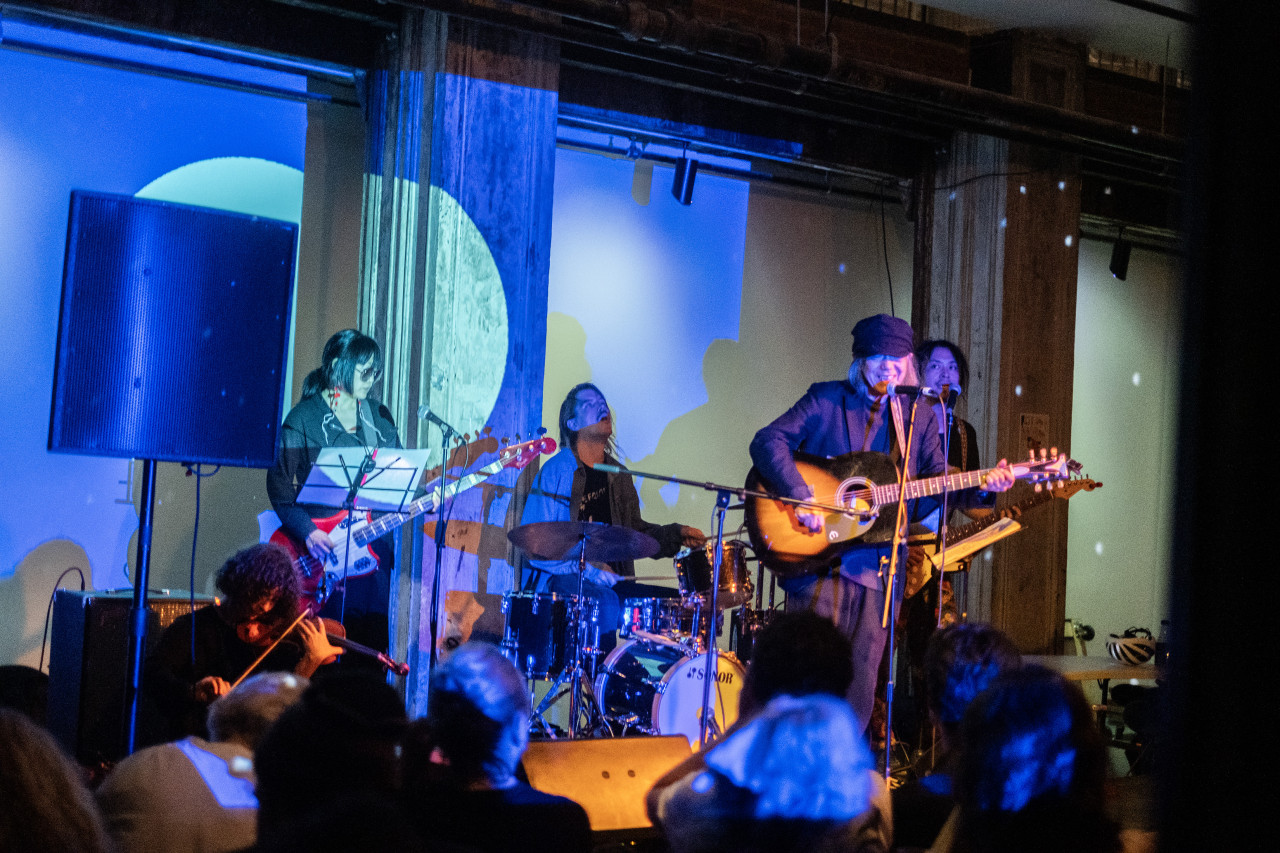 Color image of five performers on stage. They are playing bass, violin, guitars, and drums. A row of audience members are visible in the foreground, with their backs to the camera. A blue projection covers the performers and the back wall.