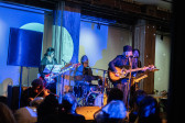 Color image of five performers on stage. They are playing bass, violin, guitars, and drums. A row of audience members are visible in the foreground, with their backs to the camera. A blue projection covers the performers and the back wall.