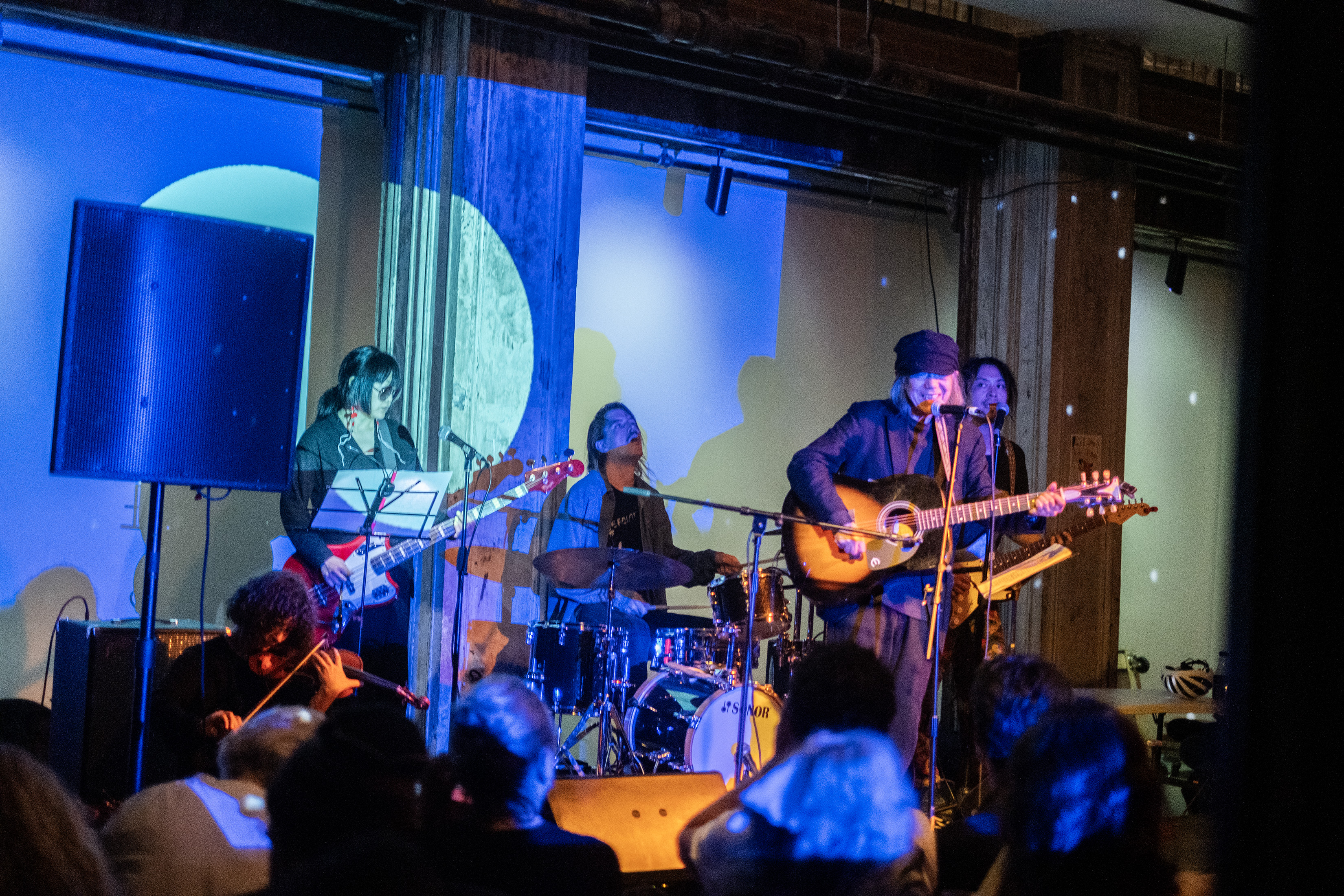 Color image of five performers on stage. They are playing bass, violin, guitars, and drums. A row of audience members are visible in the foreground, with their backs to the camera. A blue projection covers the performers and the back wall.