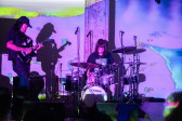 Two performers are on a stage, batehd in purple light. On the left, a performer plays the guitar and on the right, someone else plays the drums.