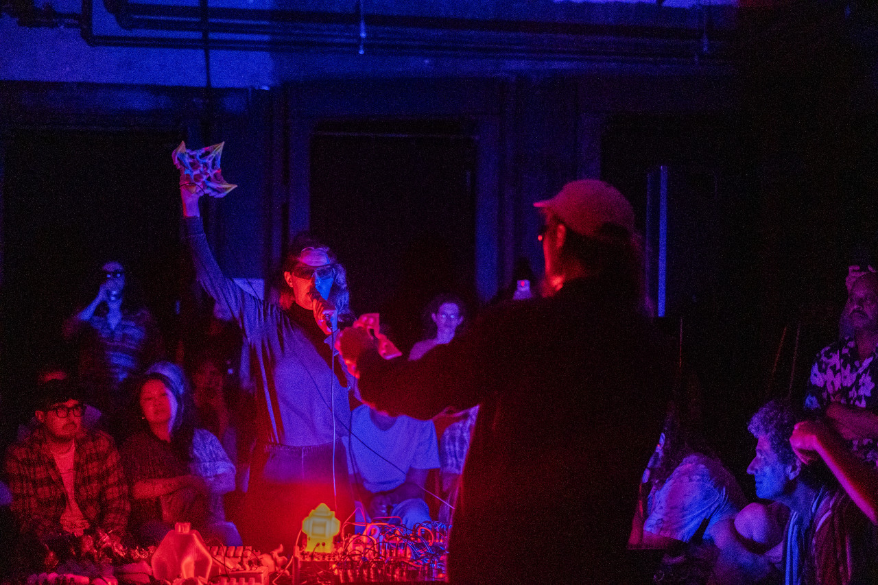 Two performers stand in the middle of a crowd, in a darkened basement. A red light casts a glow over the performers from their audio set-up in the middle of the room.
