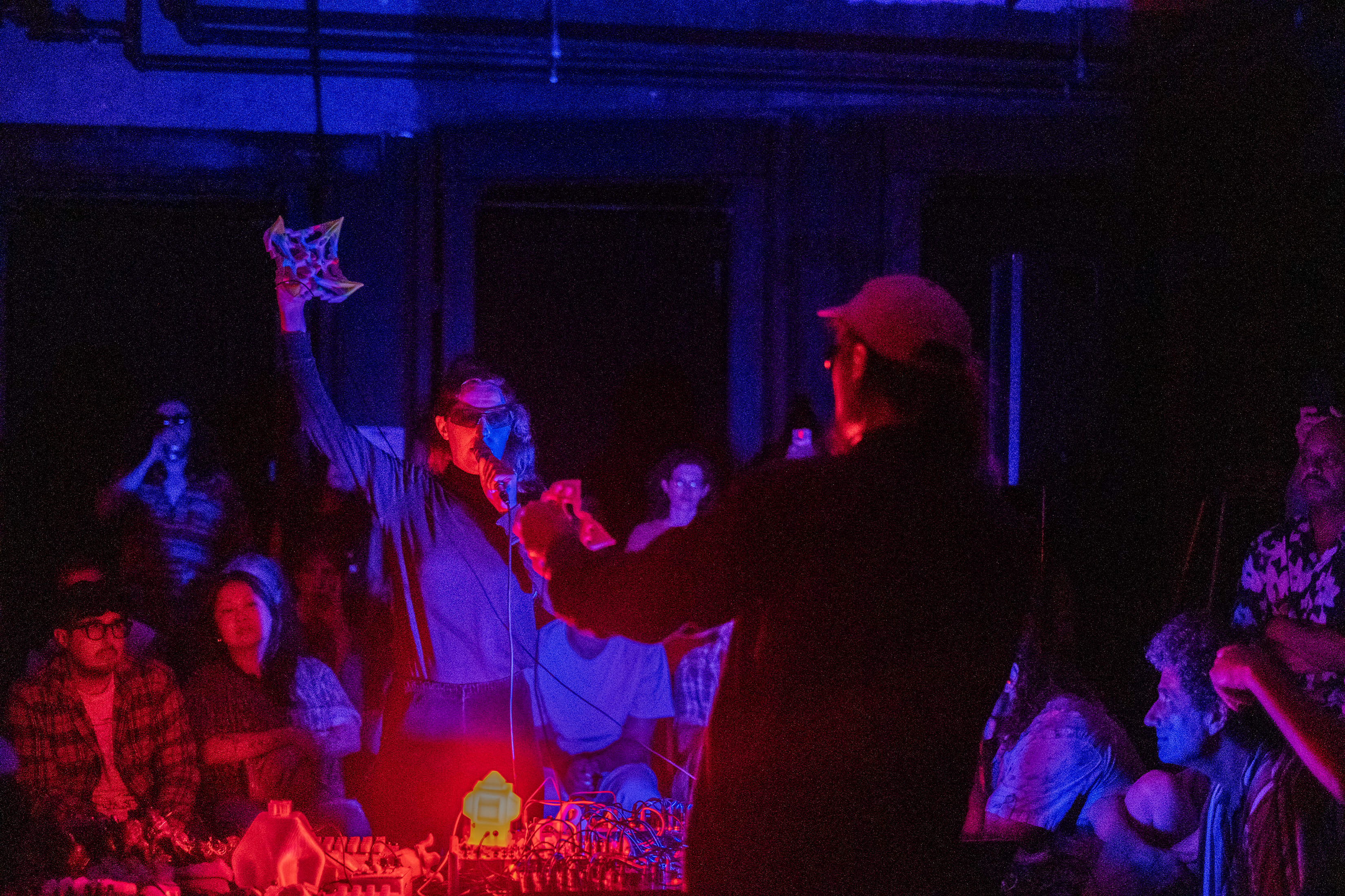 Two performers stand in the middle of a crowd, in a darkened basement. A red light casts a glow over the performers from their audio set-up in the middle of the room. 
