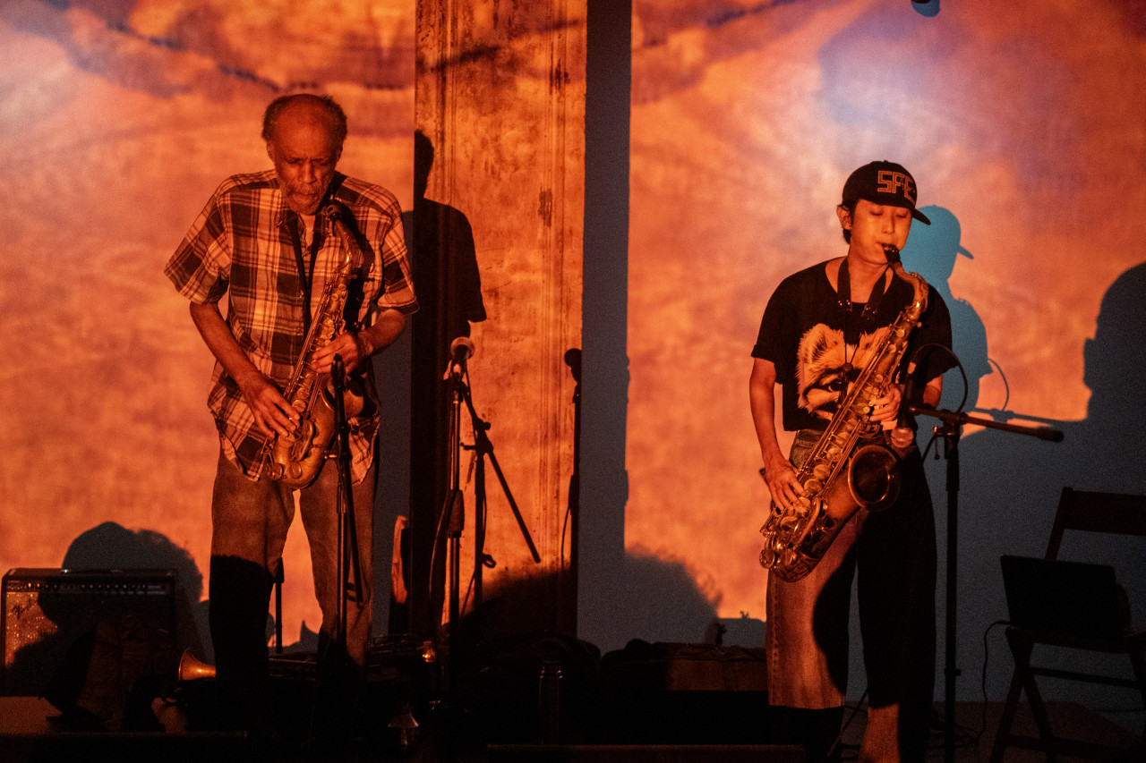 Two performers stand side by side on stage. One performer plays a brass instrument and the other a woodwind instrument. They are bathed in a red-orange light, created from a projection.