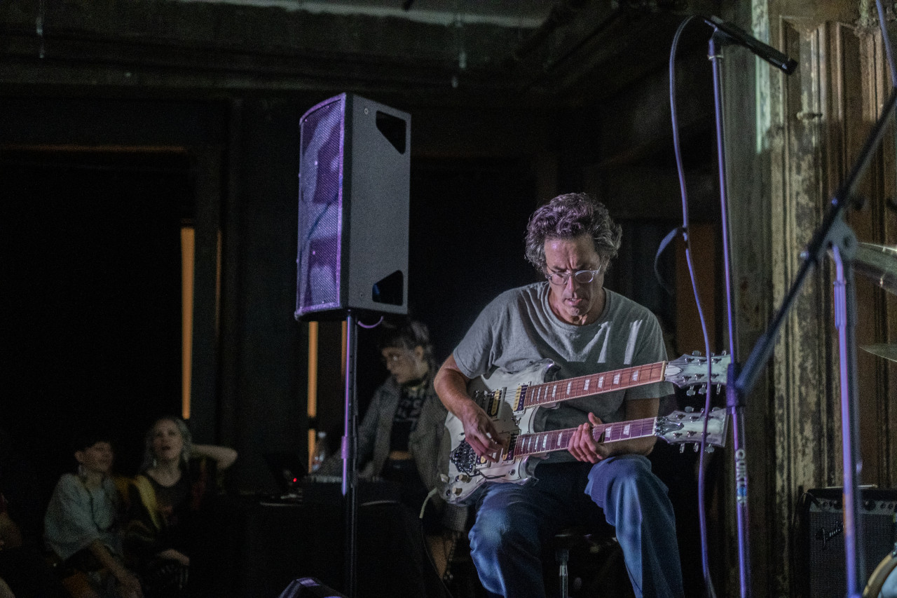 A performer sits on stage, playing a double-neck guitar.