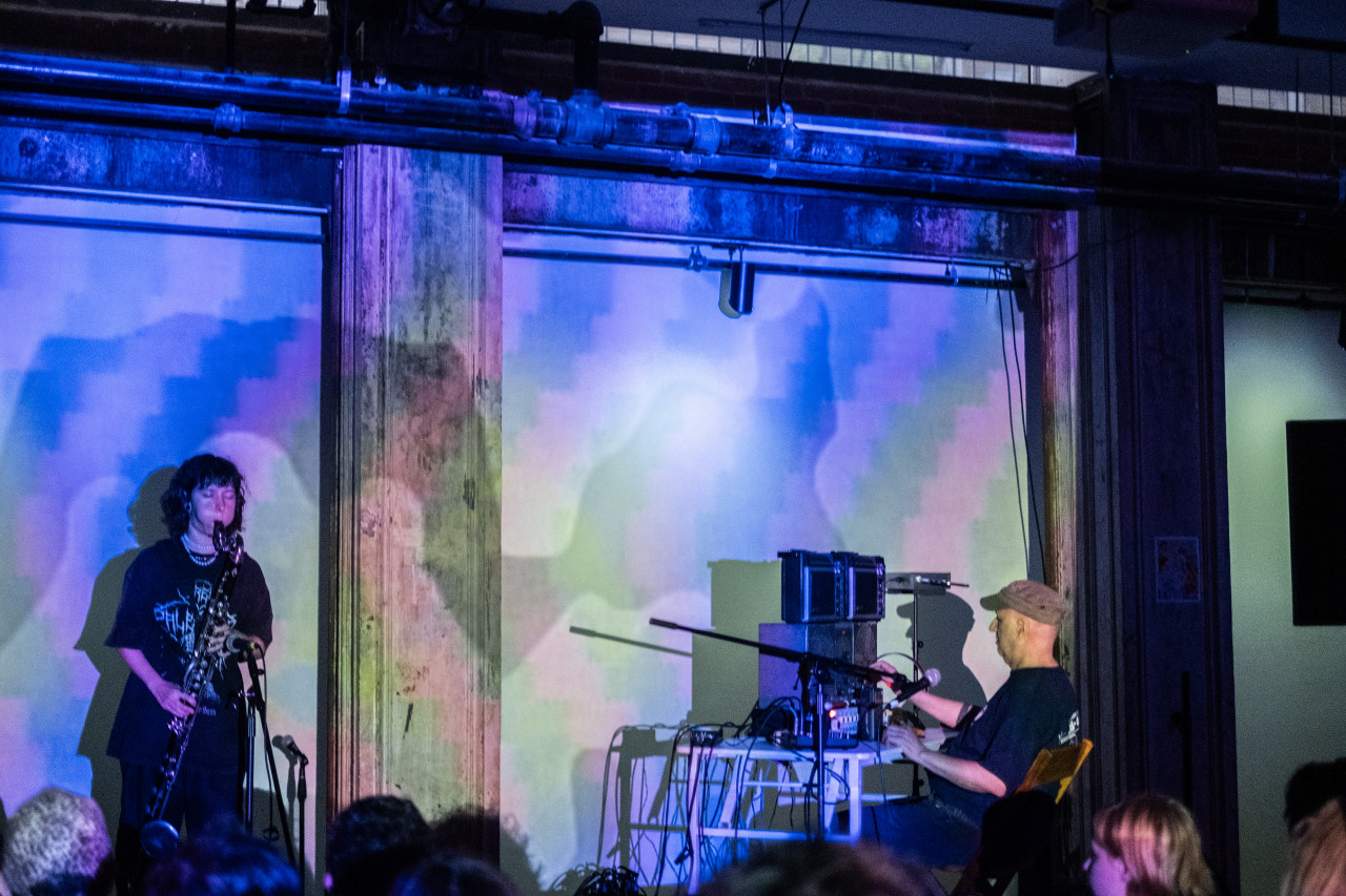 Two performers are on stage, the performer on the left side blows into a long woodwind instrument. The performer on the right sits behind a desk on top of which is a large pile of audio equipment. The stage and wall behind it are bathed in blue light, created from projections of a geometric pattern.