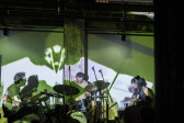 A band of three performers sit on stage. From left to right the performers play: a double-neck guitar, drums, and a bass of guitar. The room and stage is bathed in a green and white light, created from a projection.