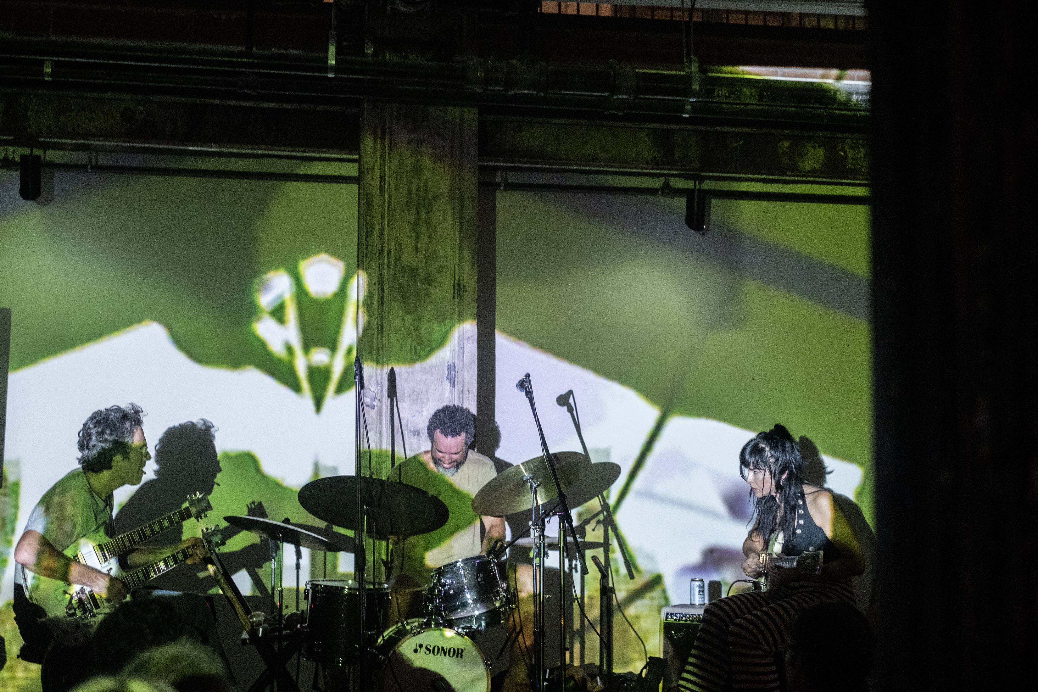 A band of three performers sit on stage. From left to right the performers play: a double-neck guitar, drums, and a bass of guitar. The room and stage is bathed in a green and white light, created from a projection.