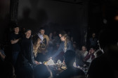 Two performers stand in the middle of a crowd in a darkened basement. A white light emanates from the table in between the performers, which also holds all of their audio equipment. The performers are bent over the table, manipulating the equipment.
