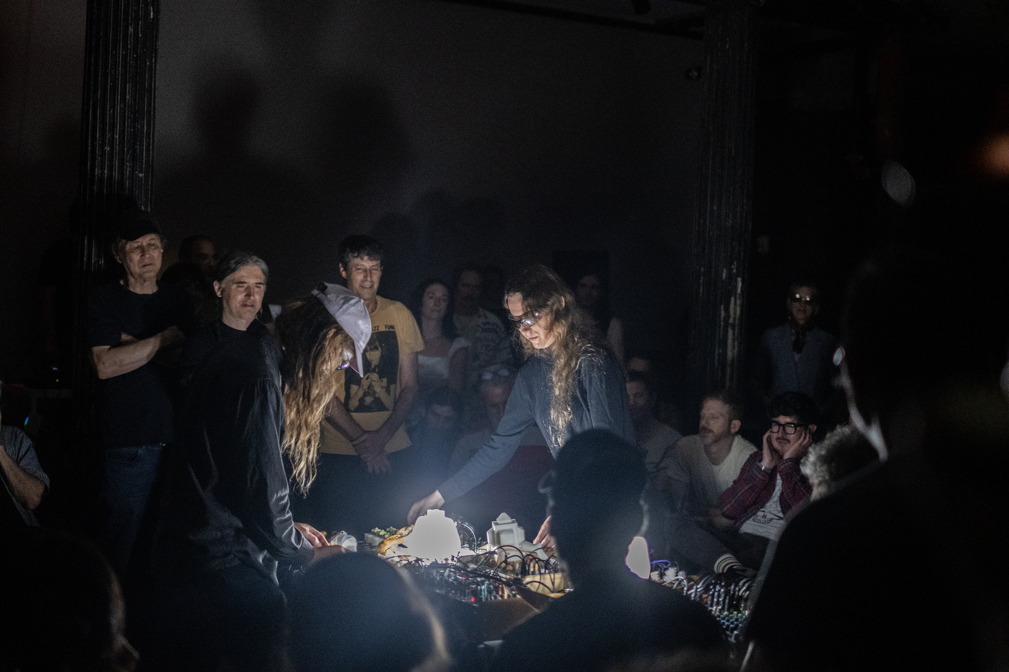 Two performers stand in the middle of a crowd in a darkened basement. A white light emanates from the table in between the performers, which also holds all of their audio equipment. The performers are bent over the table, manipulating the equipment.