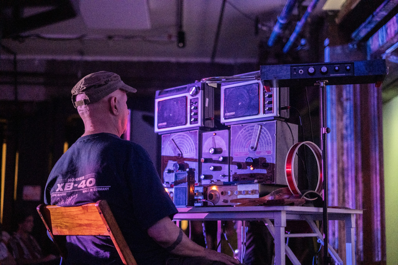 A performer sits behind a white desk, on top of which are piled various audio equipment.
