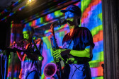 Two performers stand side by side on stage. One performer plays a brass instrument and the other a woodwind instrument. The performers and room are bathed in colorful light, created from a projection of a geometric pattern of green, blue, and red stripes.