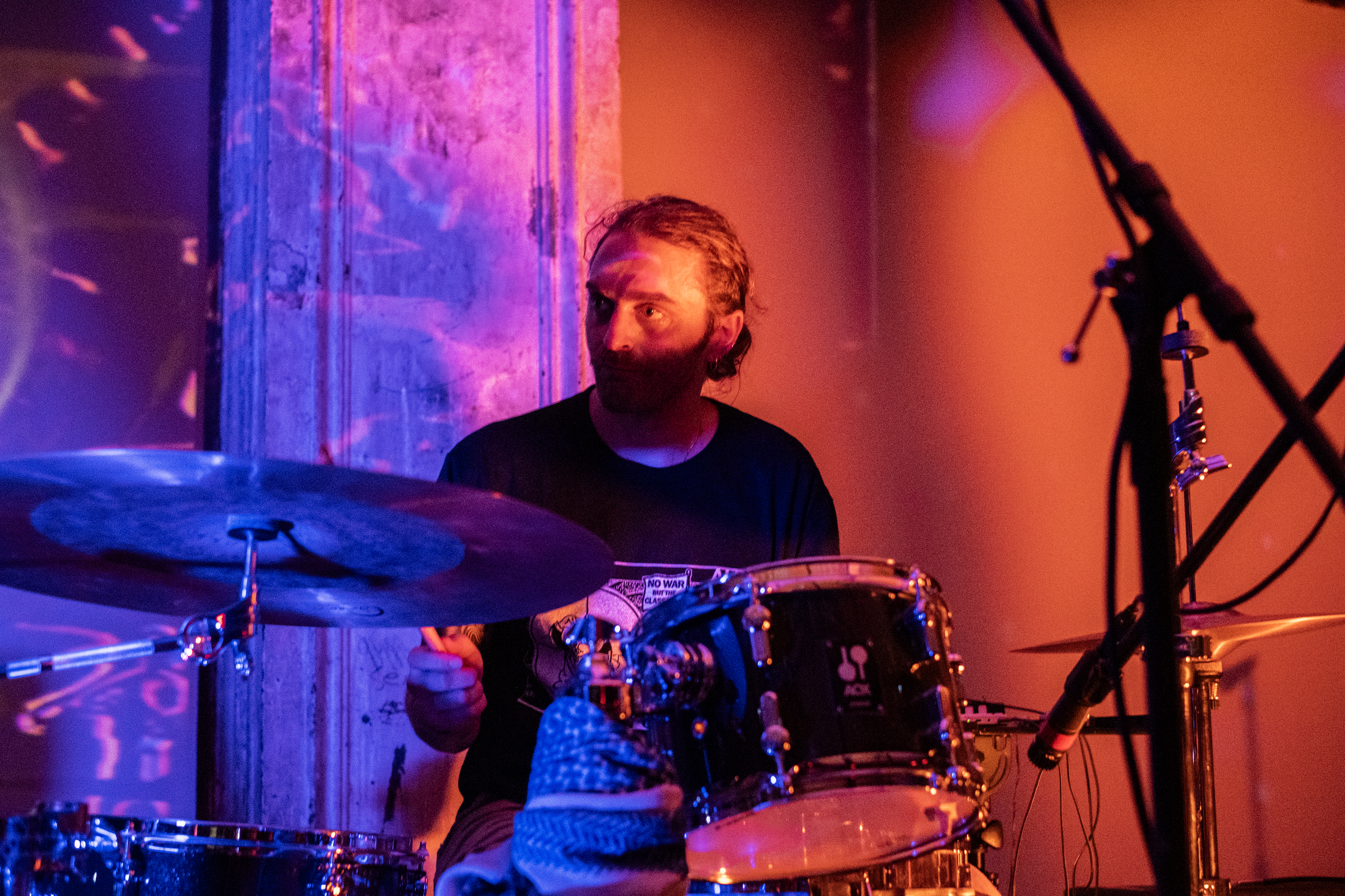 A performer plays the drums on stage. The stage and the wall behind it are bathed in blue, pink, and orange light. 