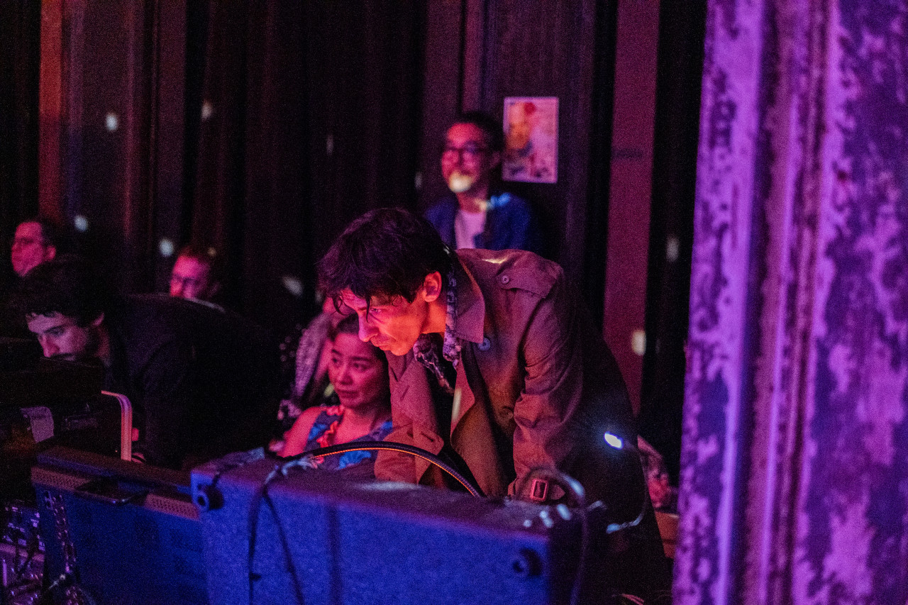 A person bathed in pink light bends over a table with equipment controlling projection visuals.