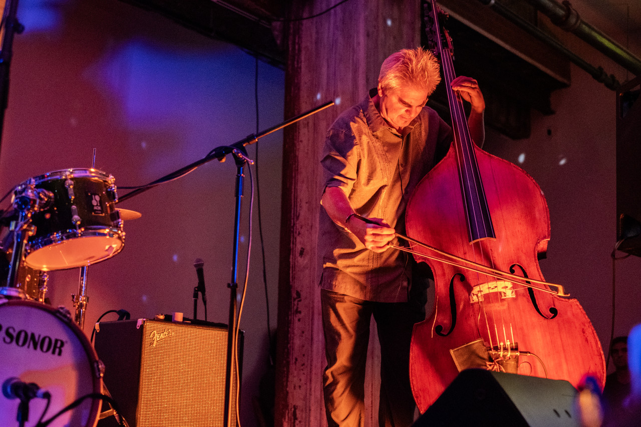 A performer plays the upright bass on stage. The stage and wall behind it are bathed in orange and blue light.