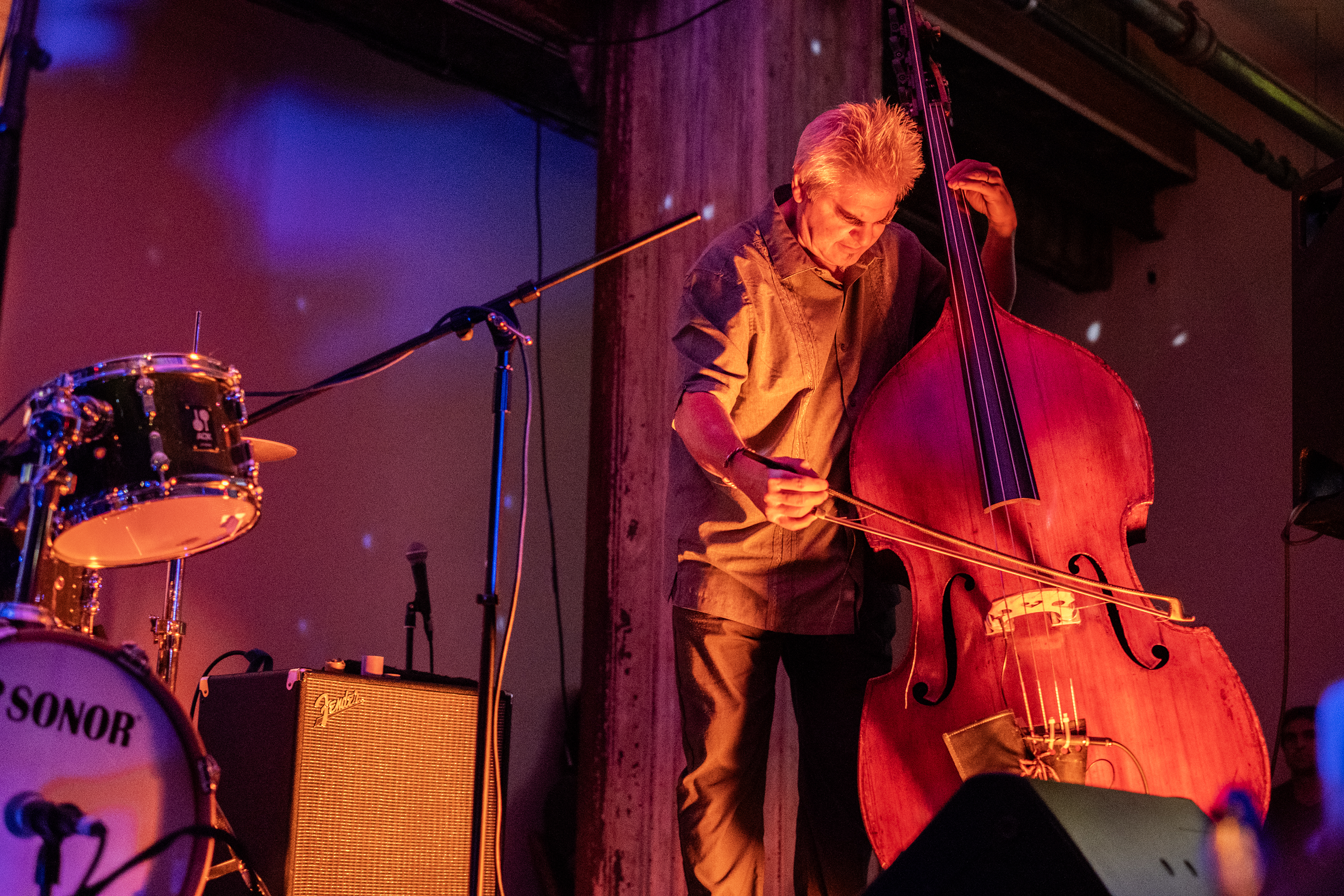 A performer plays the upright bass on stage. The stage and wall behind it are bathed in orange and blue light.