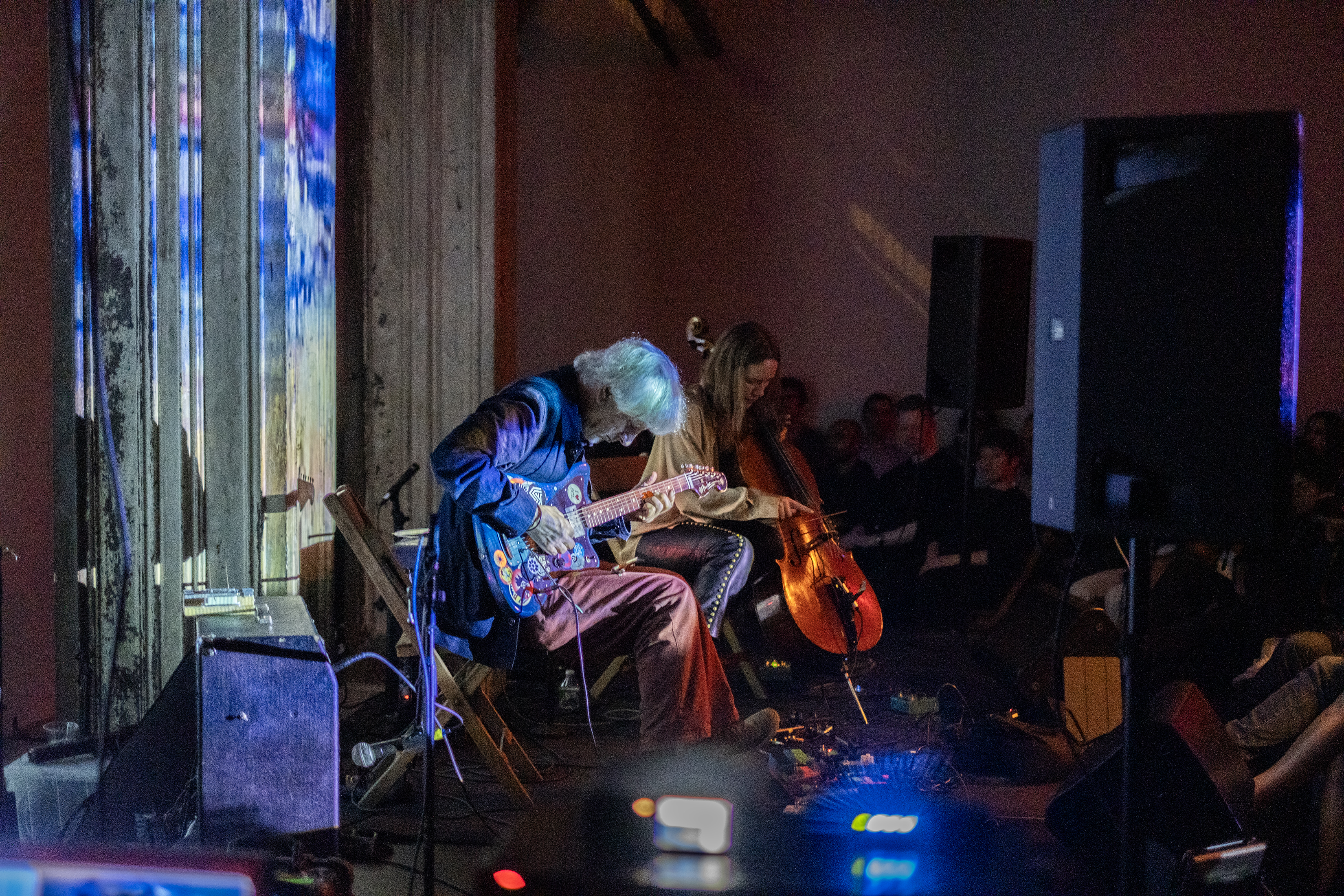 Two performers are on stage, the performer on the left side plays a guitar, and the performer on the right plays a cello. The stage and wall behind it are bathed in blue light, created from projections of a geometric pattern.
