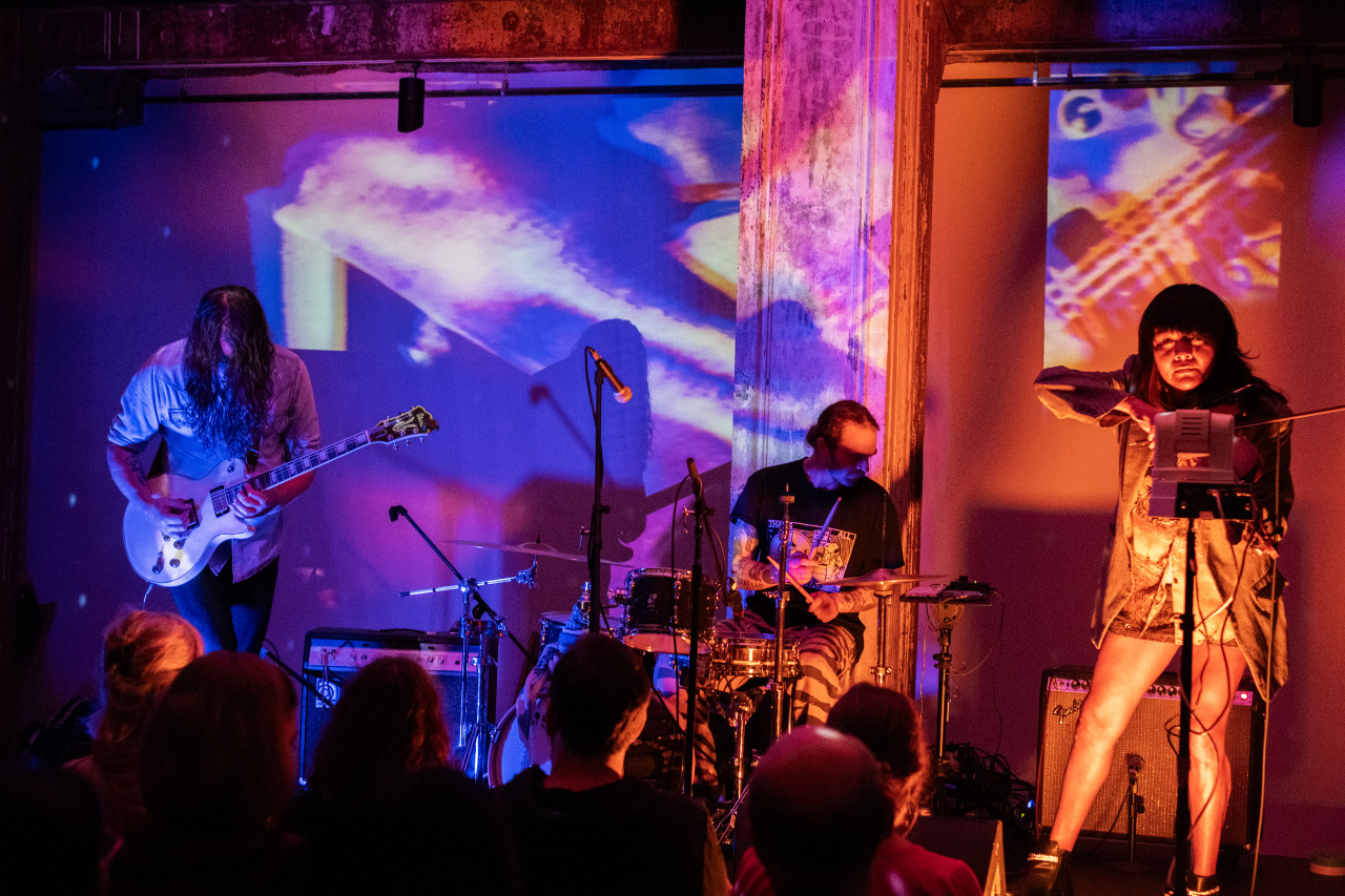 A band of three performers plays on stage. From left to right, the performers play: a guitar, drums, and a violin. The room and stage are bathed in blue, pink, and orange light created by a live video projection of the performance.