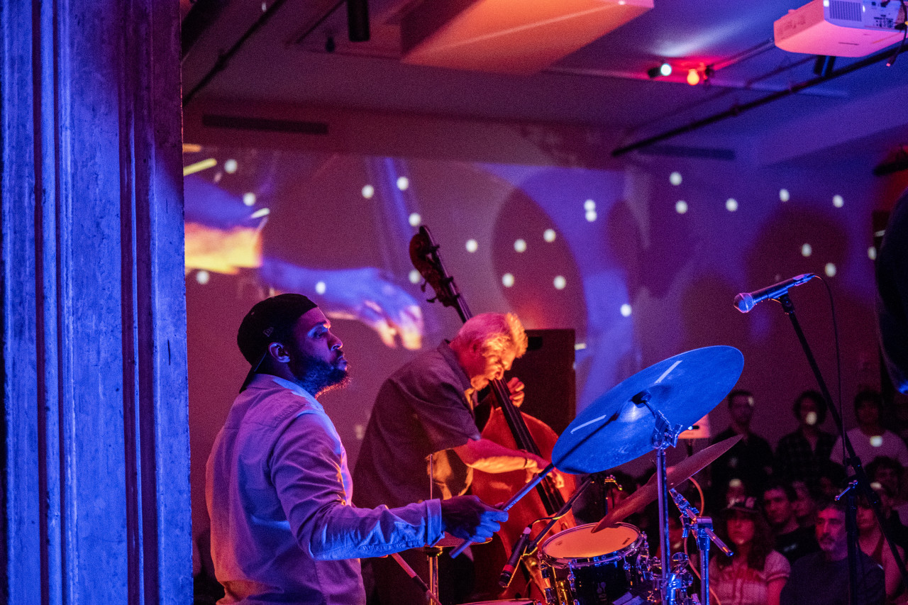 Two performers play side by side on stage. One performer plays the drums and the other a standing bass. The performers are bathed in colorful lights, created by a live video projection of the performance.