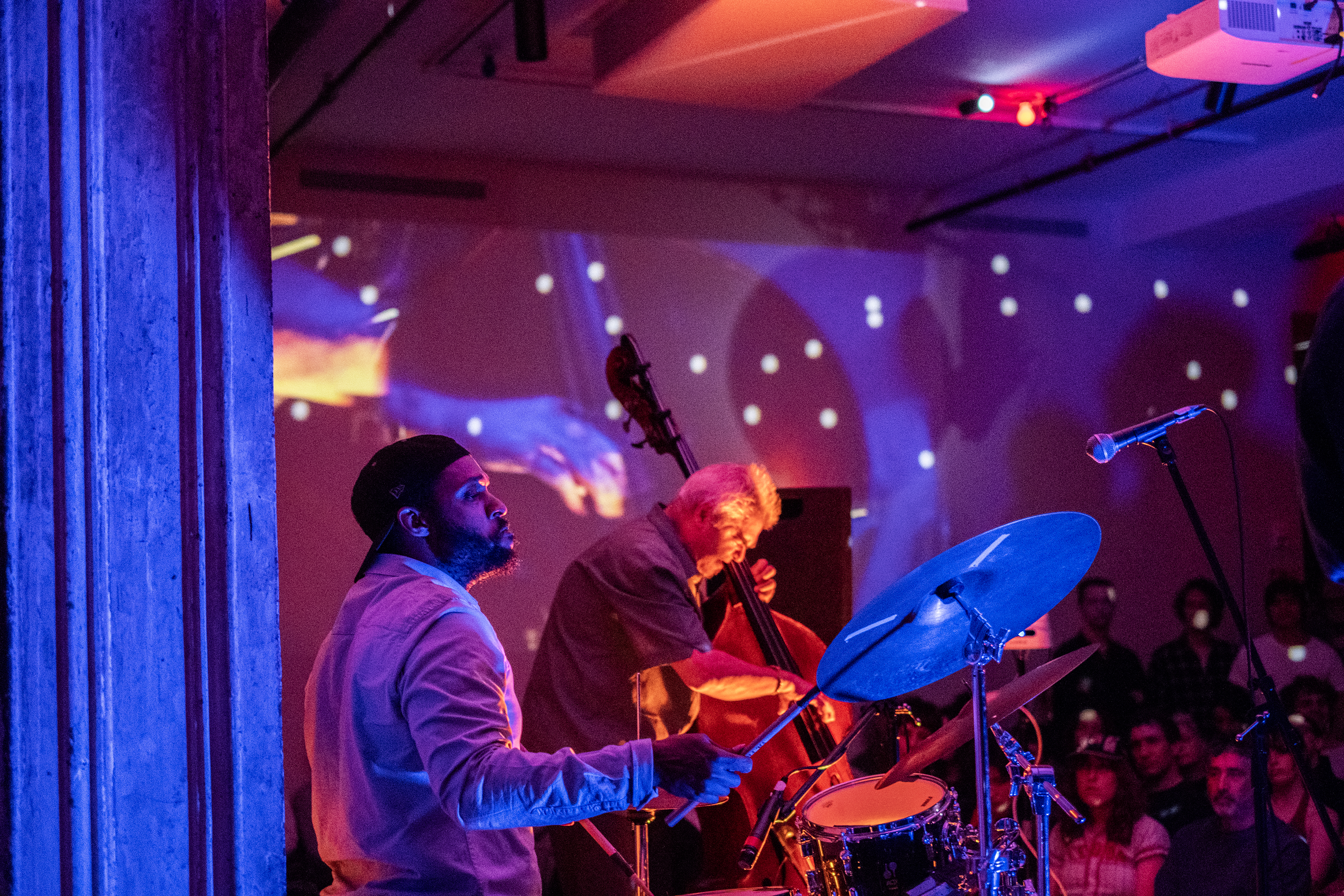 Two performers play side by side on stage. One performer plays the drums and the other a standing bass. The performers are bathed in colorful lights, created by a live video projection of the performance.