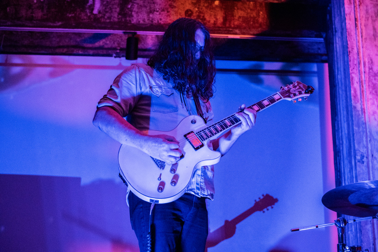 A performer plays a stringed instrument on stage. The performer and stage are bathed in pink and blue light.