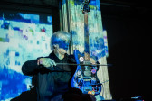 A performer plays a stringed instrument with a bow. The stage and wall behind it are bathed in blue light, created from projections of a geometric pattern.
