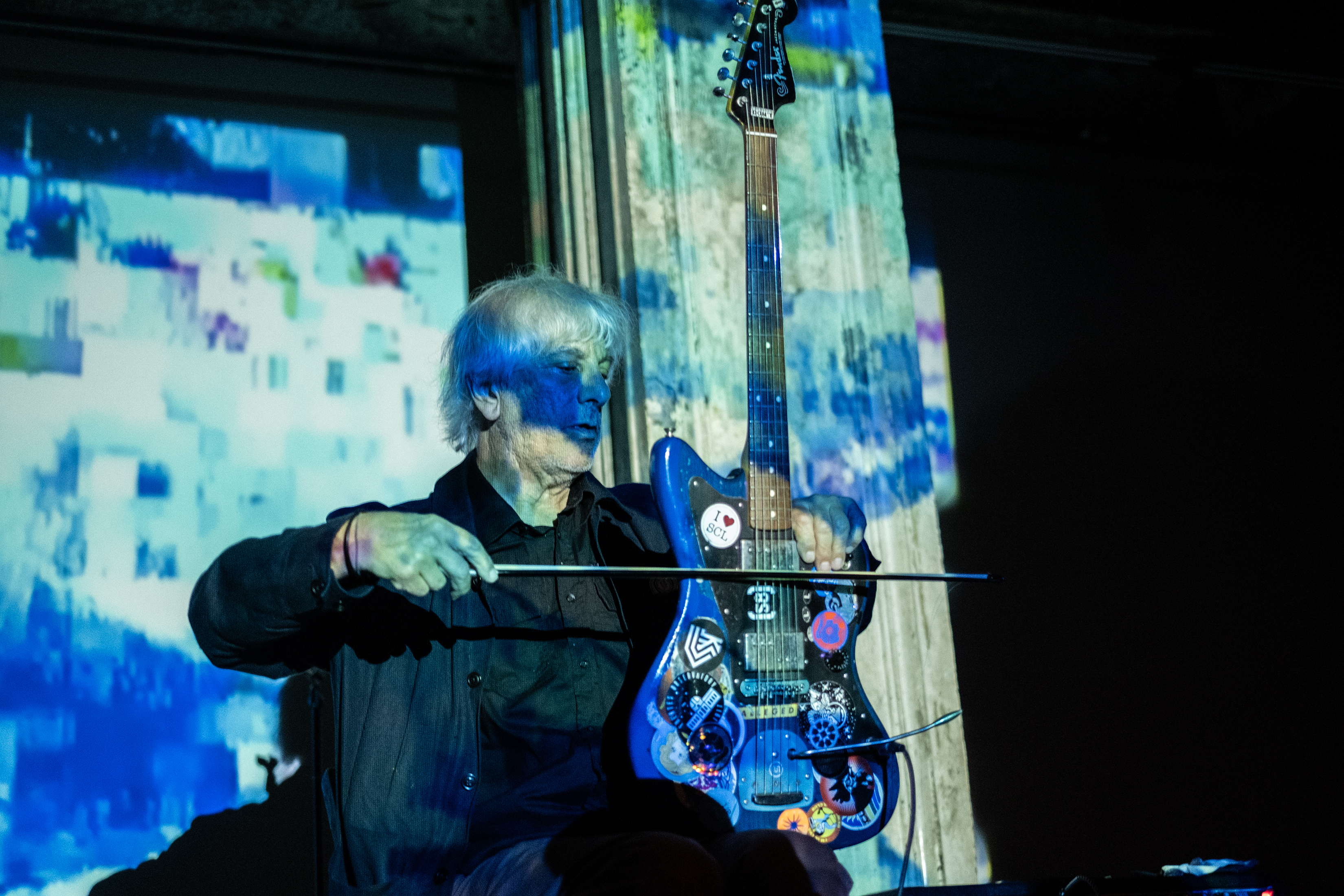 A performer plays a stringed instrument with a bow. The stage and wall behind it are bathed in blue light, created from projections of a geometric pattern.