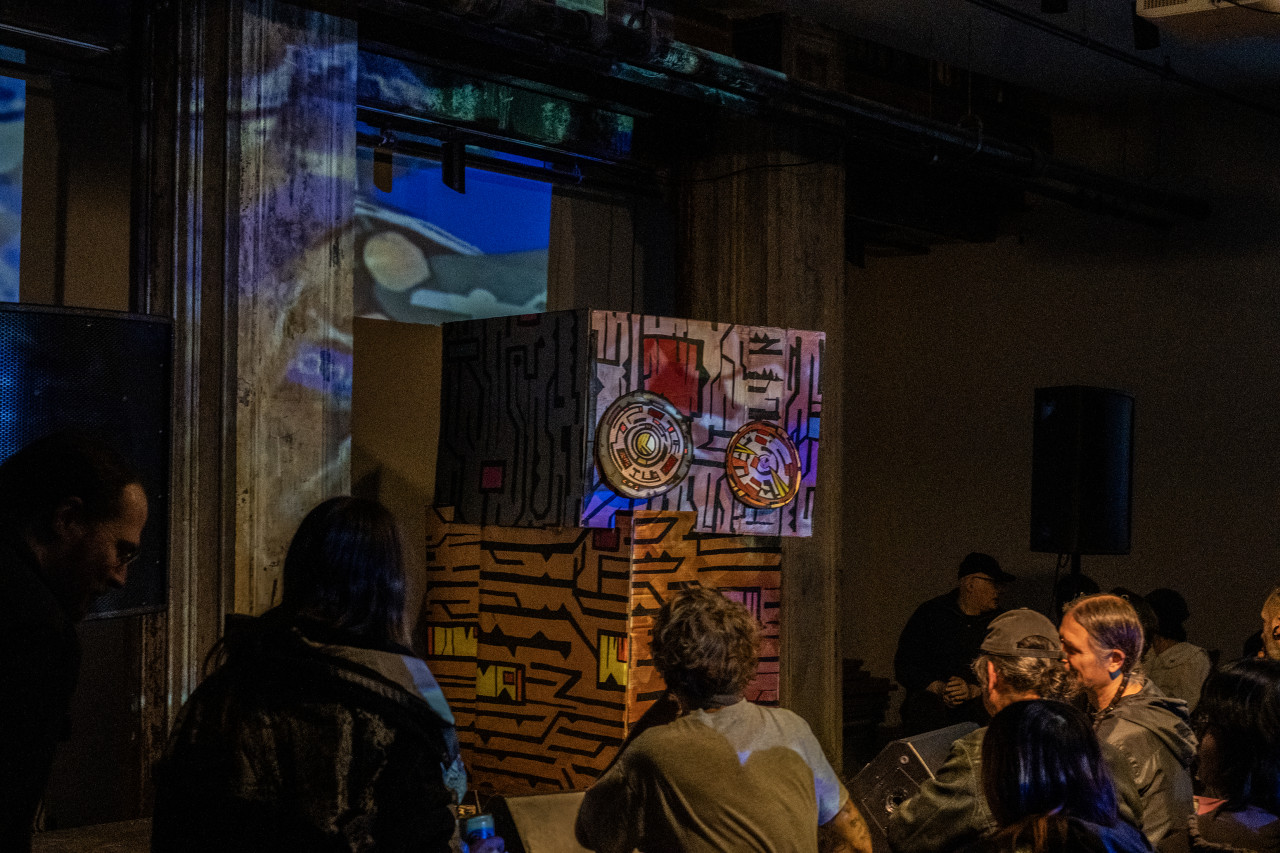 A painted cardboard structure sits on stage. The audience, stage, and wall behind it are bathed in yellow, blue, and green light created by a patterned projection.