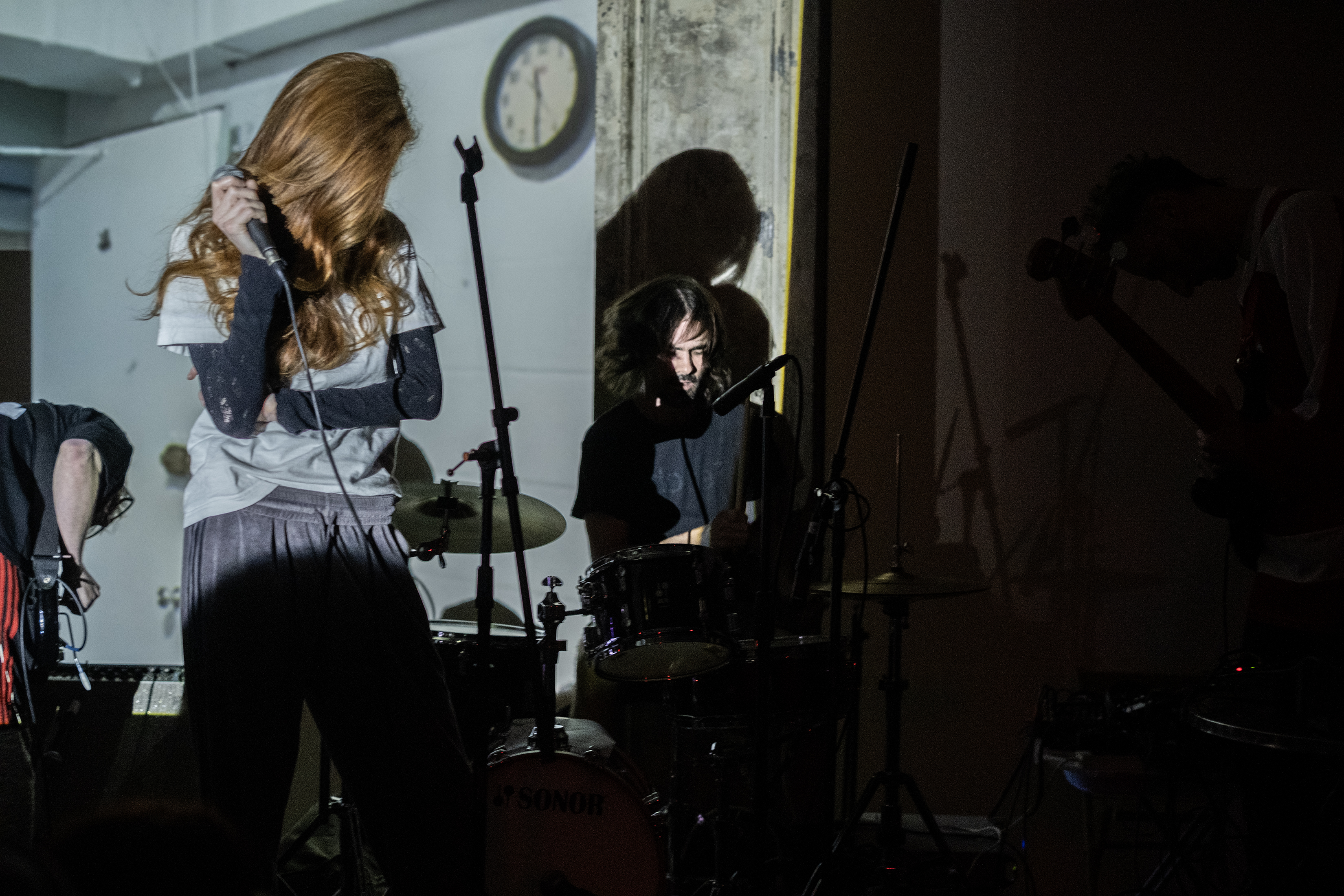 A band performs on stage, bathed in white light. The guitarist and drummer play alongside a singer, performing between them.